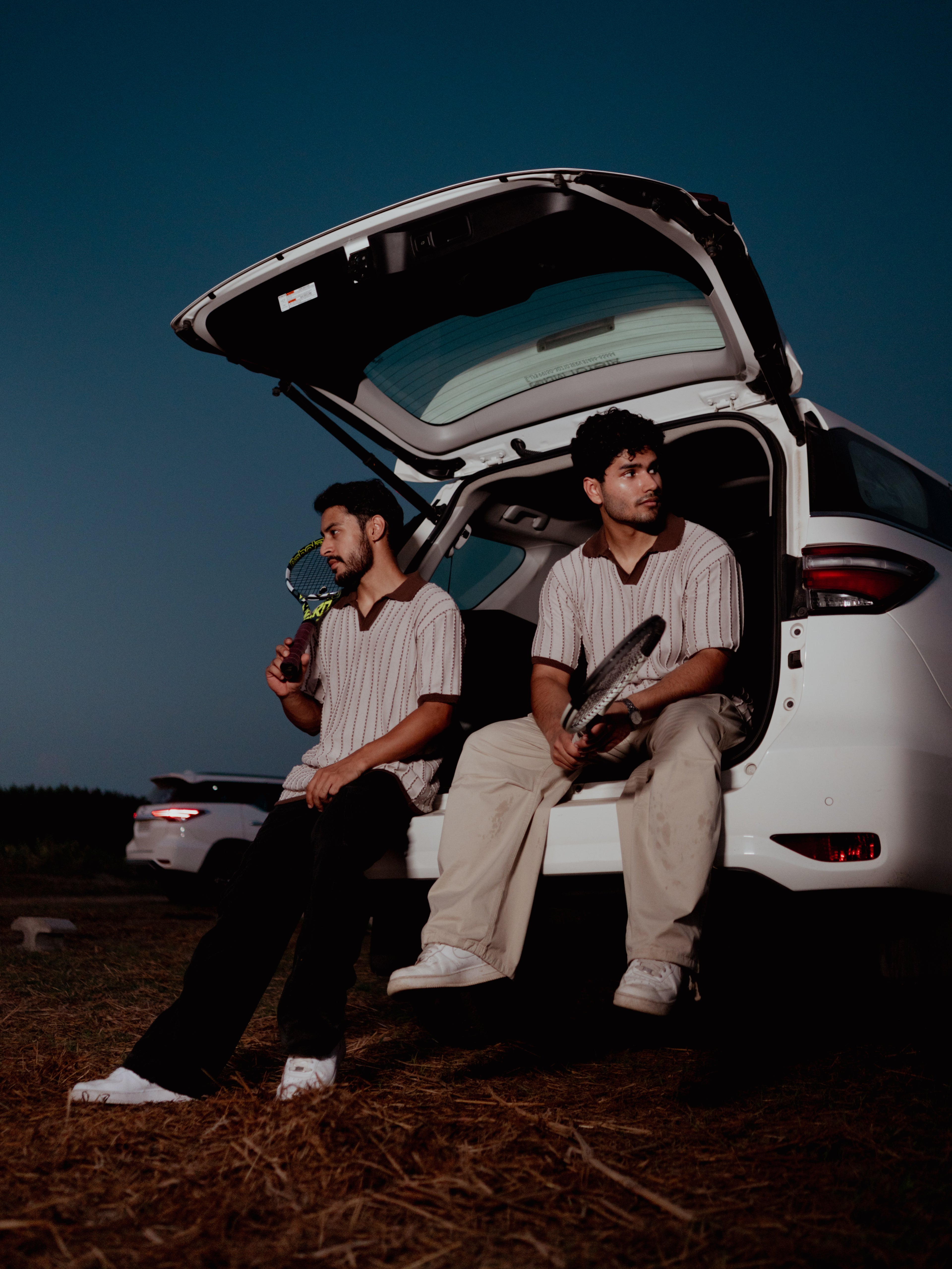 Two men sitting in the open trunk of a car at night, holding tennis rackets.